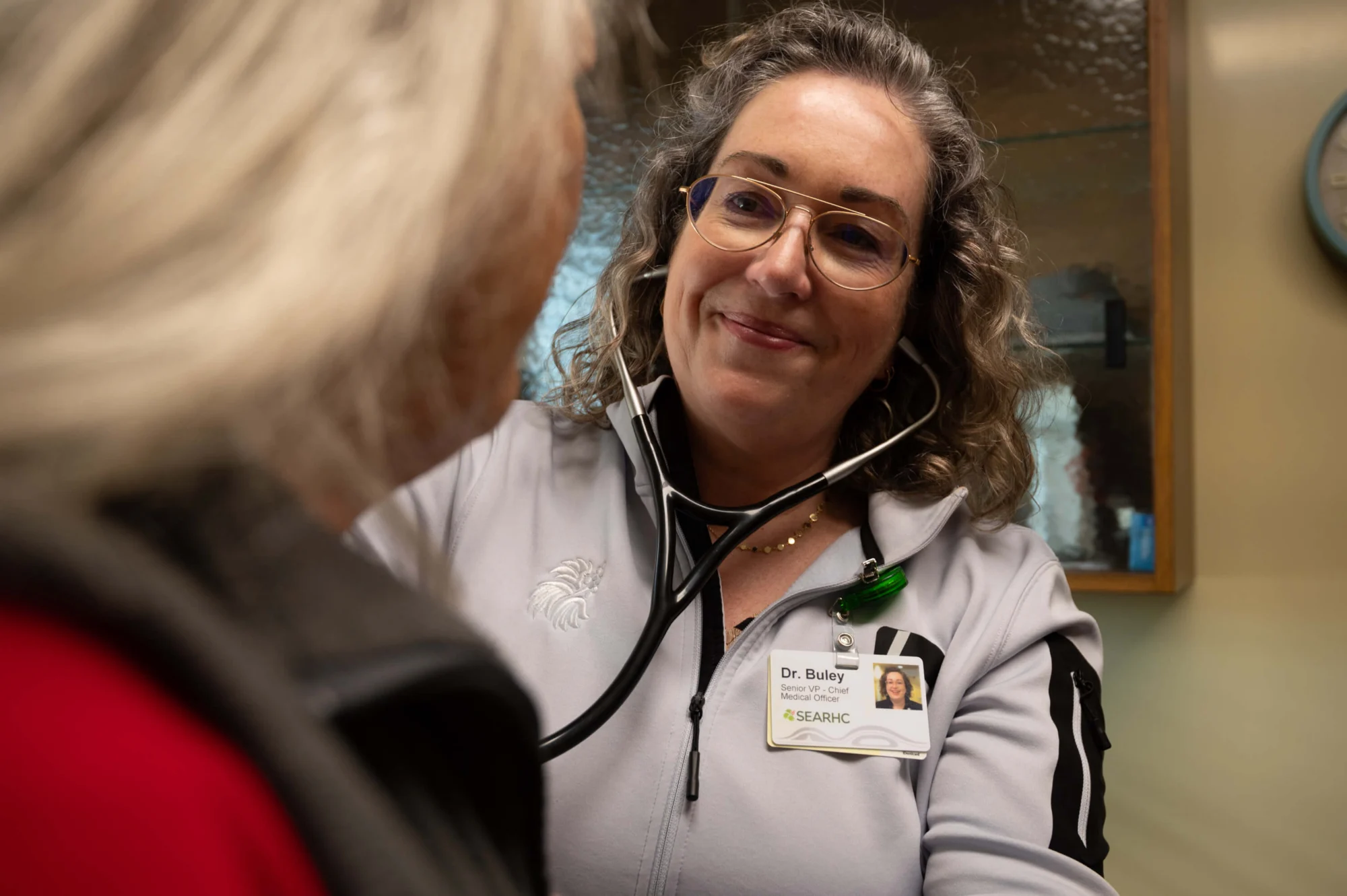 A smiling female doctor with glasses and a stethoscope around her neck looks at a patient, whose back of the head is visible. Her ID badge reads 'Dr. Buley, Senior VP - Chief Medical Officer, SEARHC'.