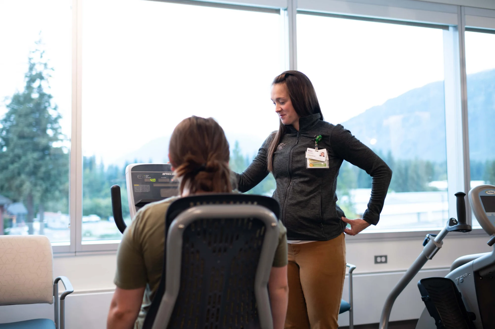 A physical therapist guides a patient on a recumbent exercise bike in a room with large windows overlooking mountains and trees.