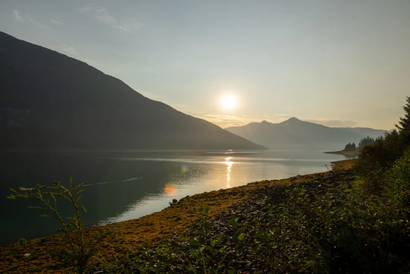 Sunrise or sunset over a mountain lake, with the sun reflecting golden light on the water, surrounded by dark mountains and a forested shoreline.