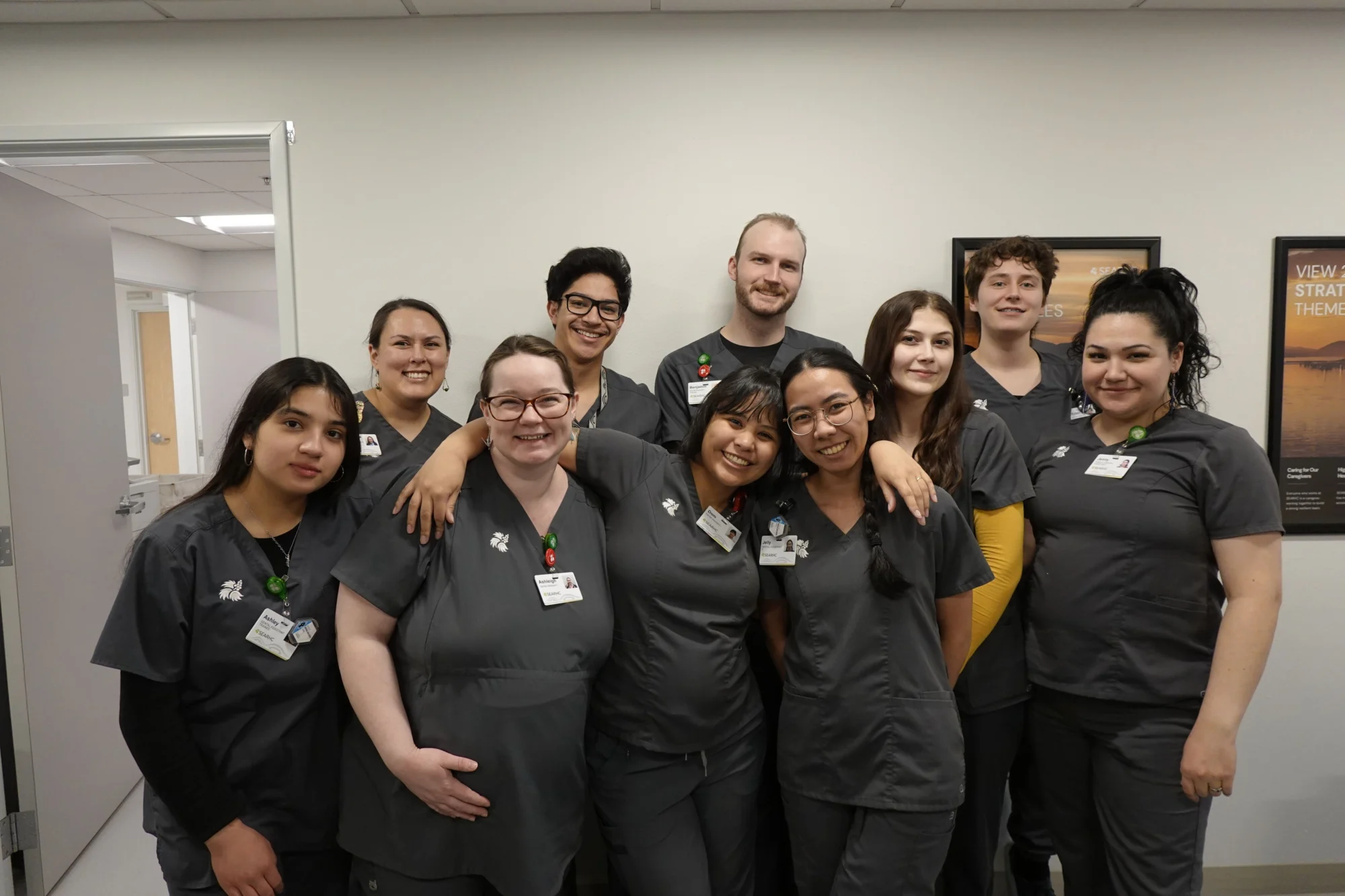 Ten smiling healthcare professionals in dark gray scrubs pose for a group photo indoors. They wear SEARHC ID badges, and some have arms around each other.