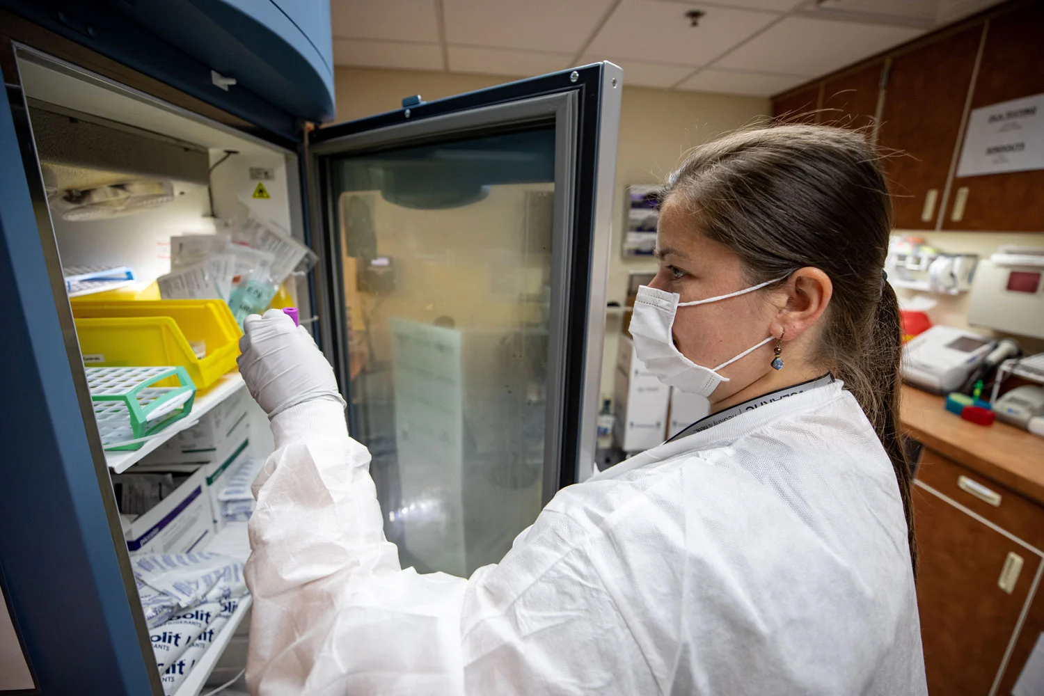 A person in a lab coat, mask, and gloves reaches into an open lab refrigerator, holding a purple-capped tube.