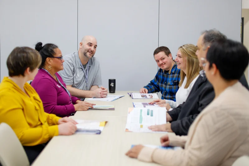 Seven people sit around a table in a meeting, looking at documents and conversing. Some are smiling.