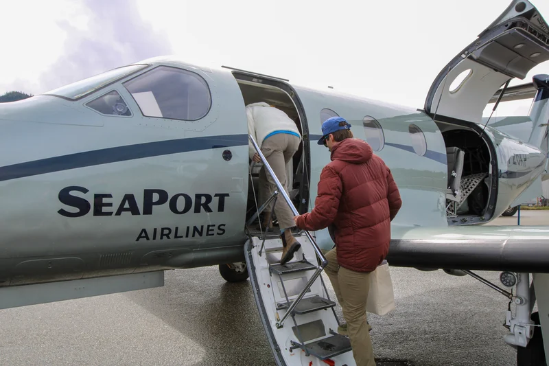 Two people boarding a light gray SeaPort Airlines propeller plane with integrated stairs on a wet tarmac.