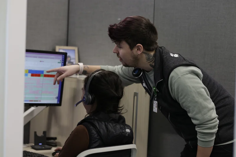 Two people in an office, one standing and pointing at a computer screen while the other is seated wearing a headset.