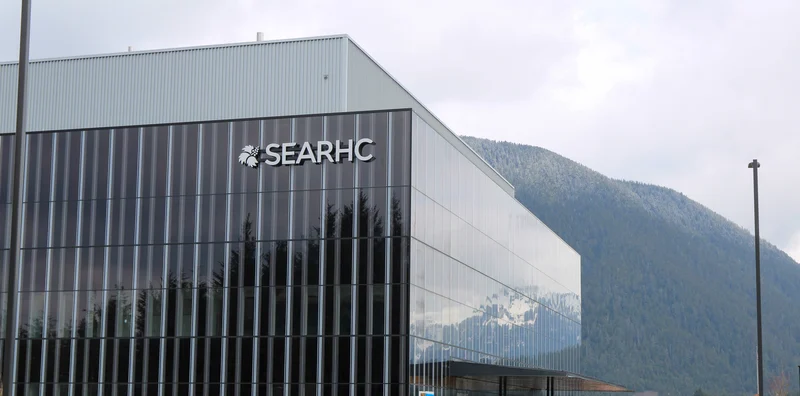 Exterior of a modern building with 'SEARHC' sign on a glass facade, with a snow-dusted mountain in the background.