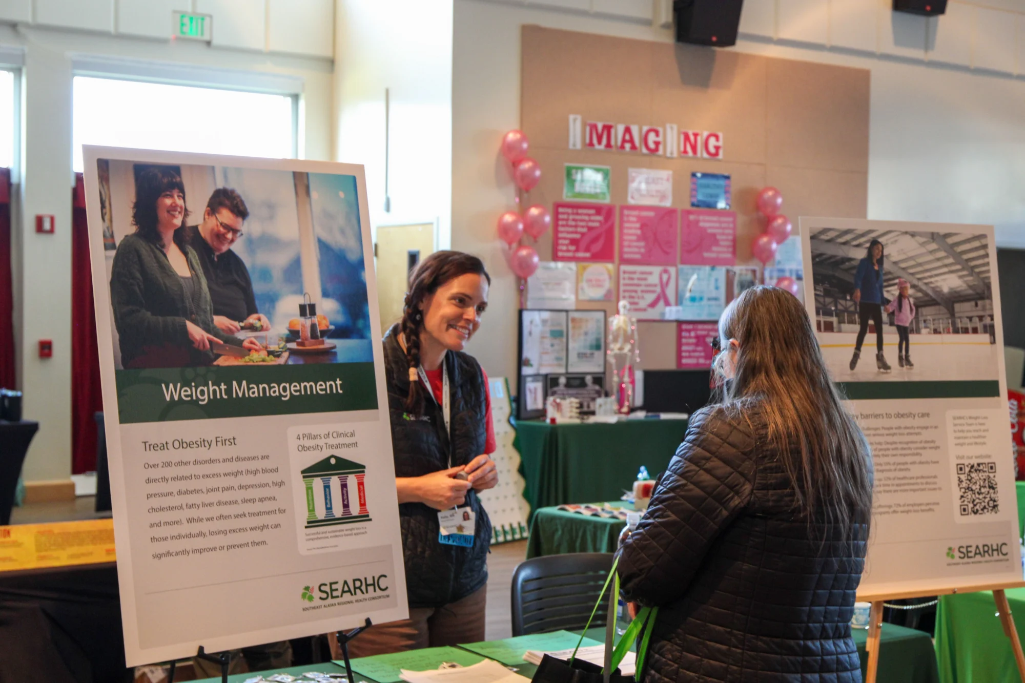 Two women at a health fair booth. One woman, facing forward, smiles while talking to another woman whose back is to the camera. Signs for 'Weight Management' and 'IMAGING' are visible.