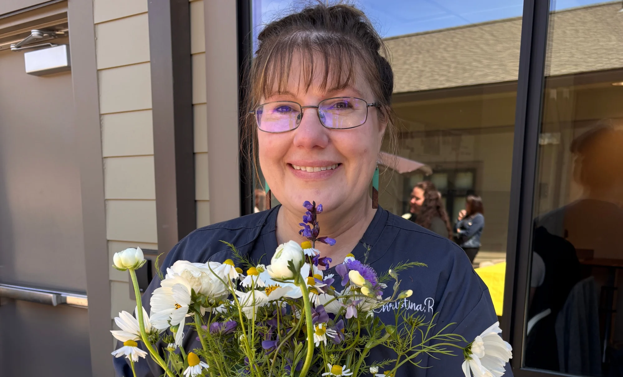 A smiling woman in glasses and a dark blue scrub top holds a bouquet of white and purple flowers.