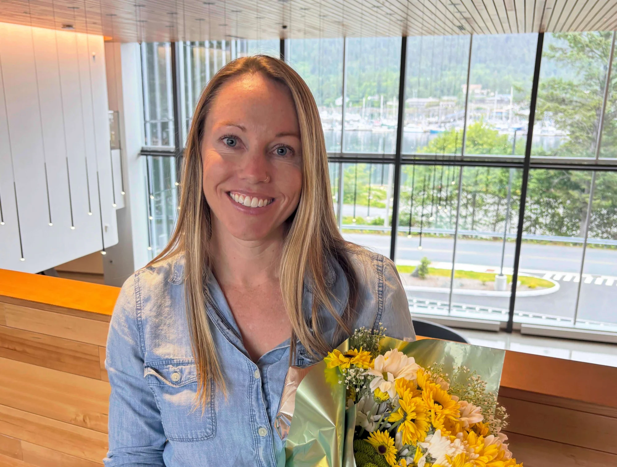 A smiling woman with blonde hair and blue eyes holds a bouquet of yellow and white flowers in front of a large window overlooking a marina and mountains.