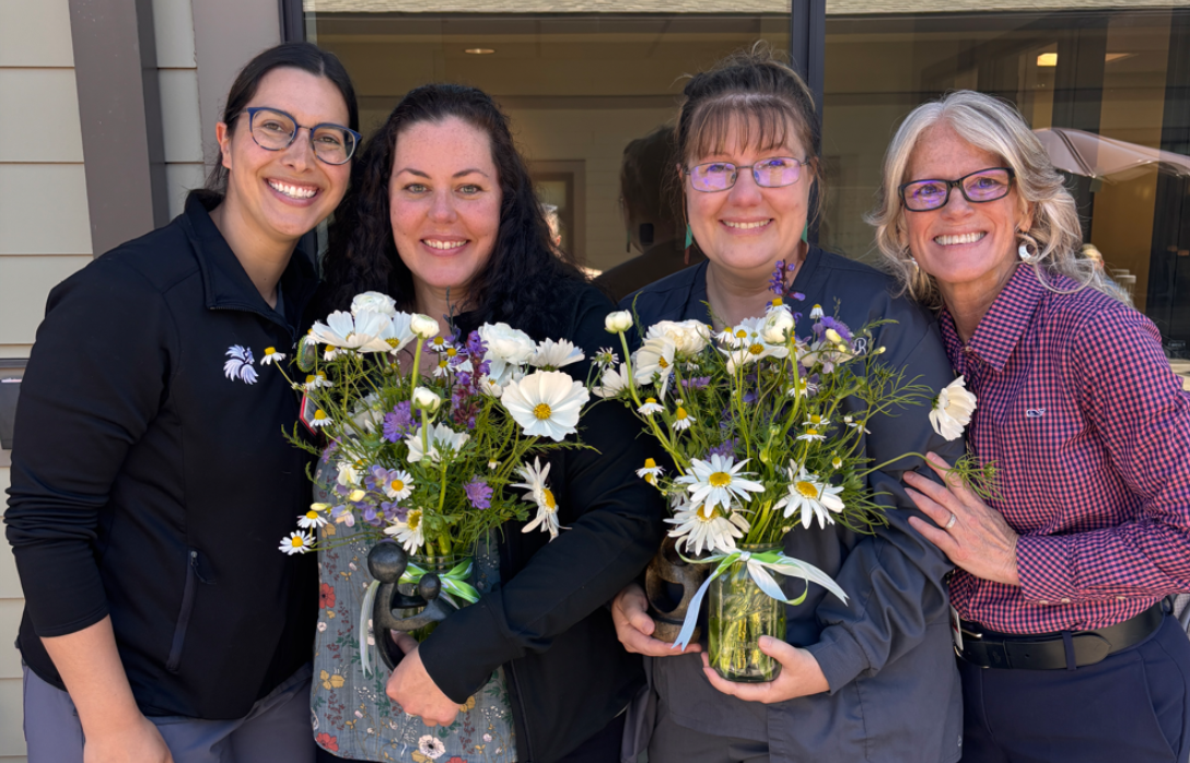 Four smiling women standing side-by-side, holding bouquets of white and purple flowers.