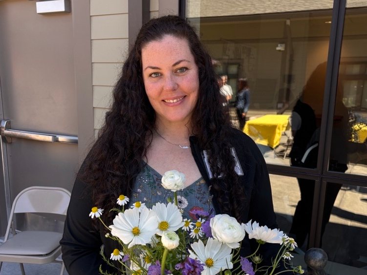 A smiling woman with long, dark, curly hair holds a bouquet of white and purple flowers.