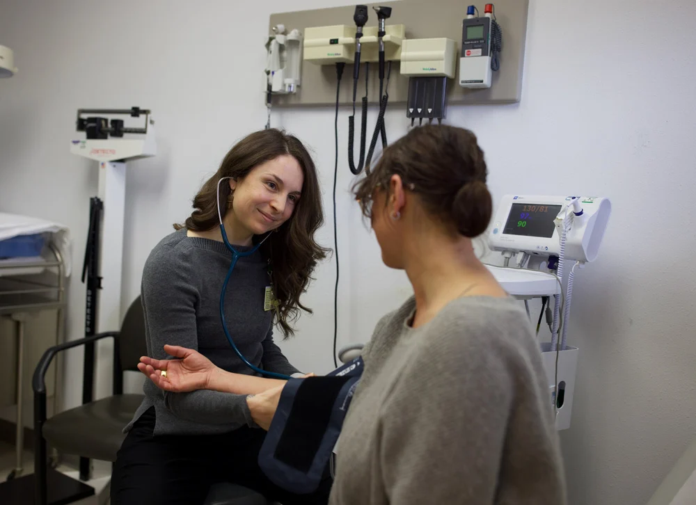 A smiling female healthcare professional with a stethoscope interacts with a patient during a blood pressure check in a medical exam room.
