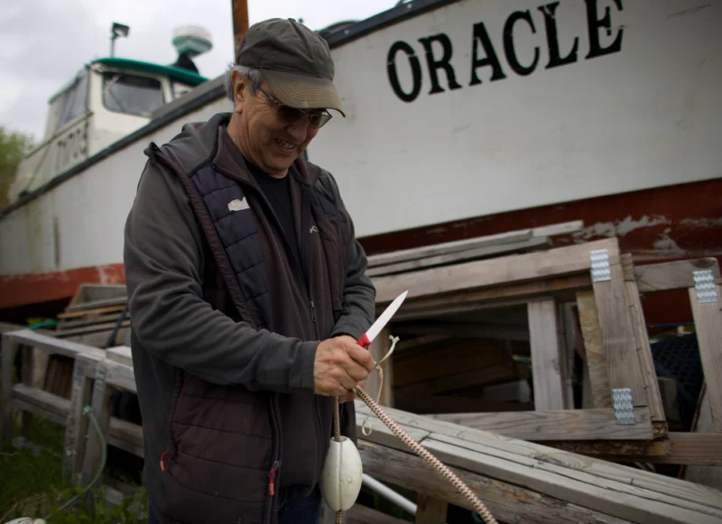 A smiling man in a dark cap and vest cuts a rope with a knife, standing in front of a white boat named "ORACLE" in a boatyard.