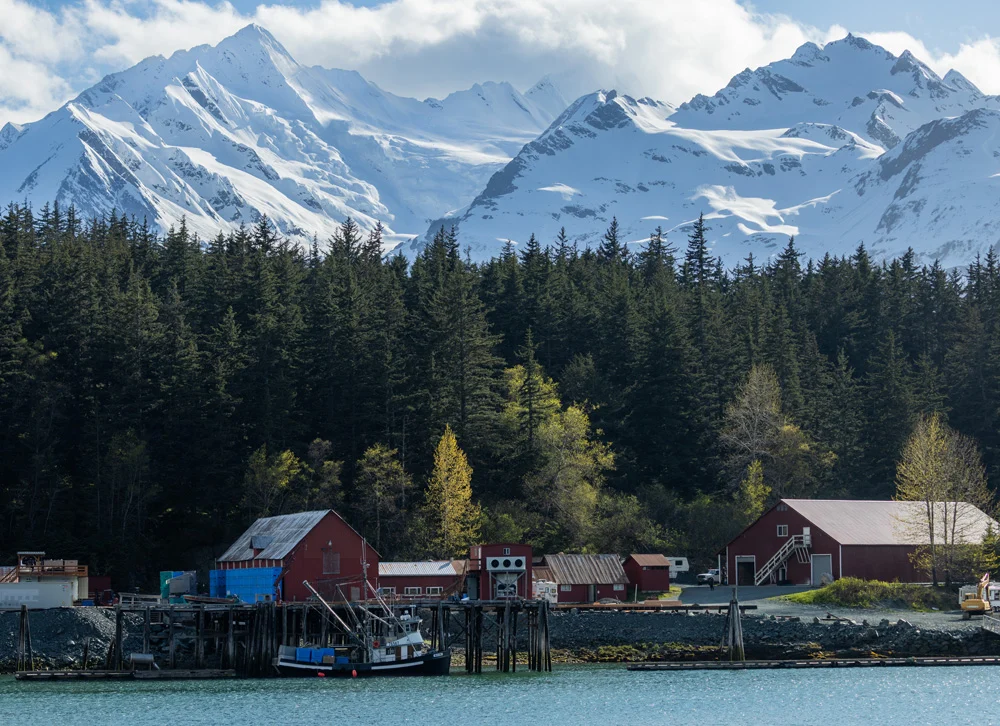 A fishing boat docked at a pier with red buildings and a dense evergreen forest behind it. Towering snow-capped mountains rise in the background under a partly cloudy sky, reflecting in the turquoise water.