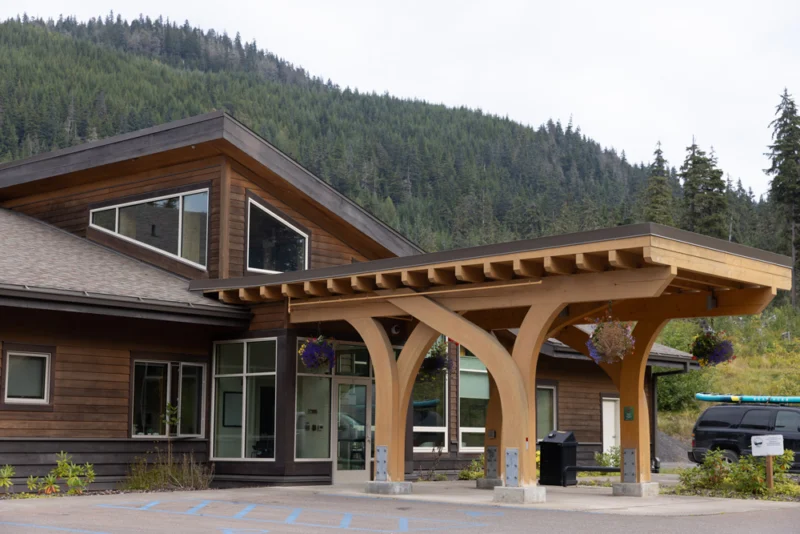 A modern wooden building with a large, light-colored timber entrance and unique windows, backed by a dense green forested mountain under an overcast sky. A black SUV with a kayak is parked nearby.