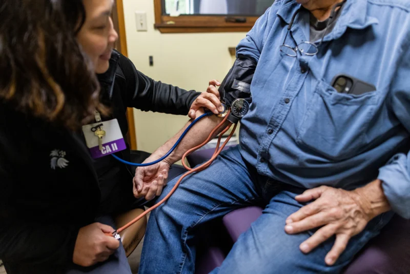 A healthcare worker with an ID badge is taking an older man's blood pressure with a cuff and stethoscope.