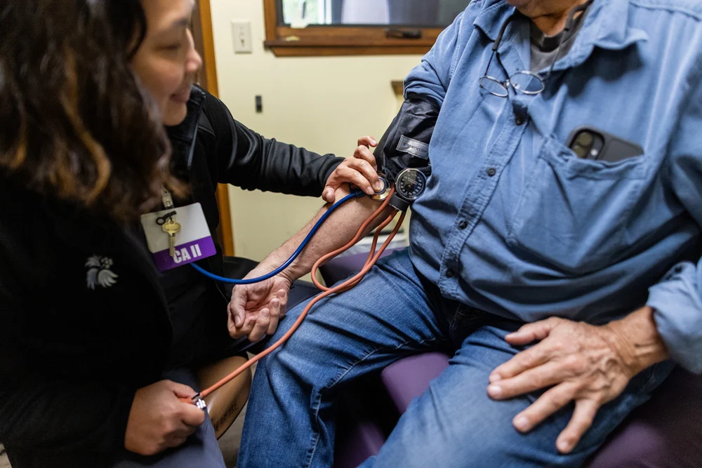 A healthcare worker with an ID badge is taking an older man's blood pressure with a cuff and stethoscope.