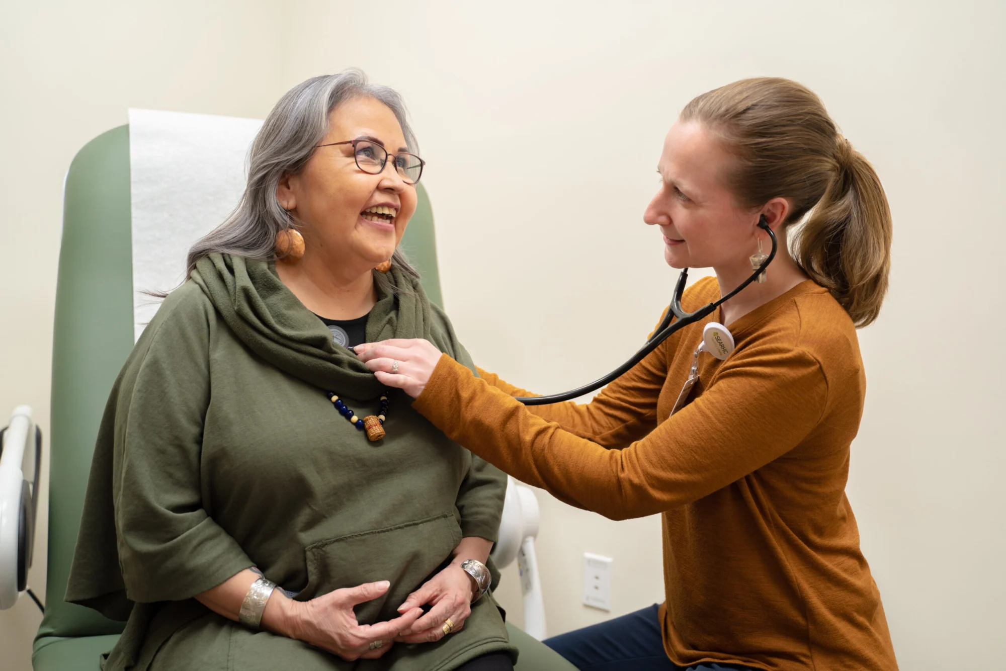 A medical professional uses a stethoscope to examine a smiling patient in a clinic.