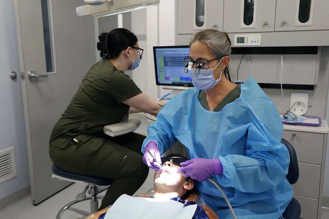 A dentist or hygienist in a blue gown and mask works on a patient's teeth, while an assistant in green scrubs uses a computer in the background.