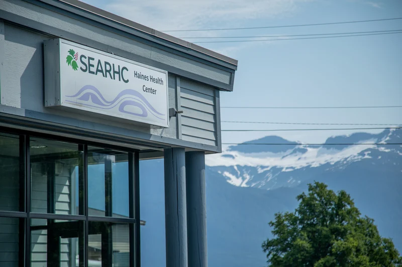 Exterior view of the SEARHC Haines Health Center building with a sign, glass windows, and snow-capped mountains in the background.