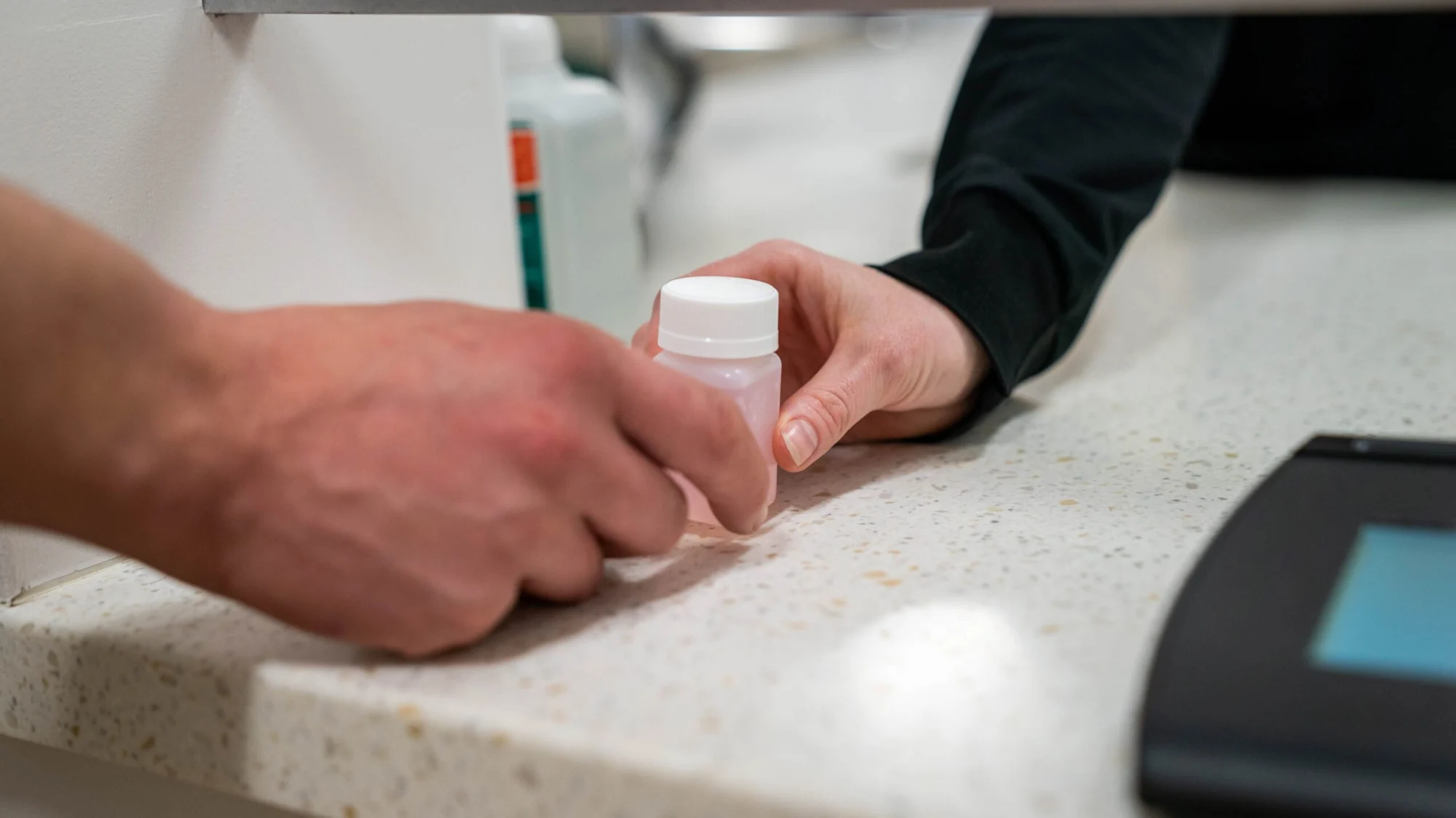 Two hands exchanging a small pink bottle with a white cap over a speckled countertop.