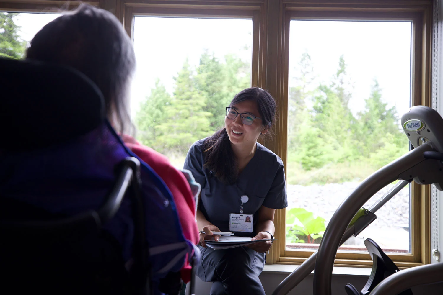 A smiling healthcare professional in scrubs sits on a window sill, talking to a patient in a wheelchair, with a green forest visible outside the window.