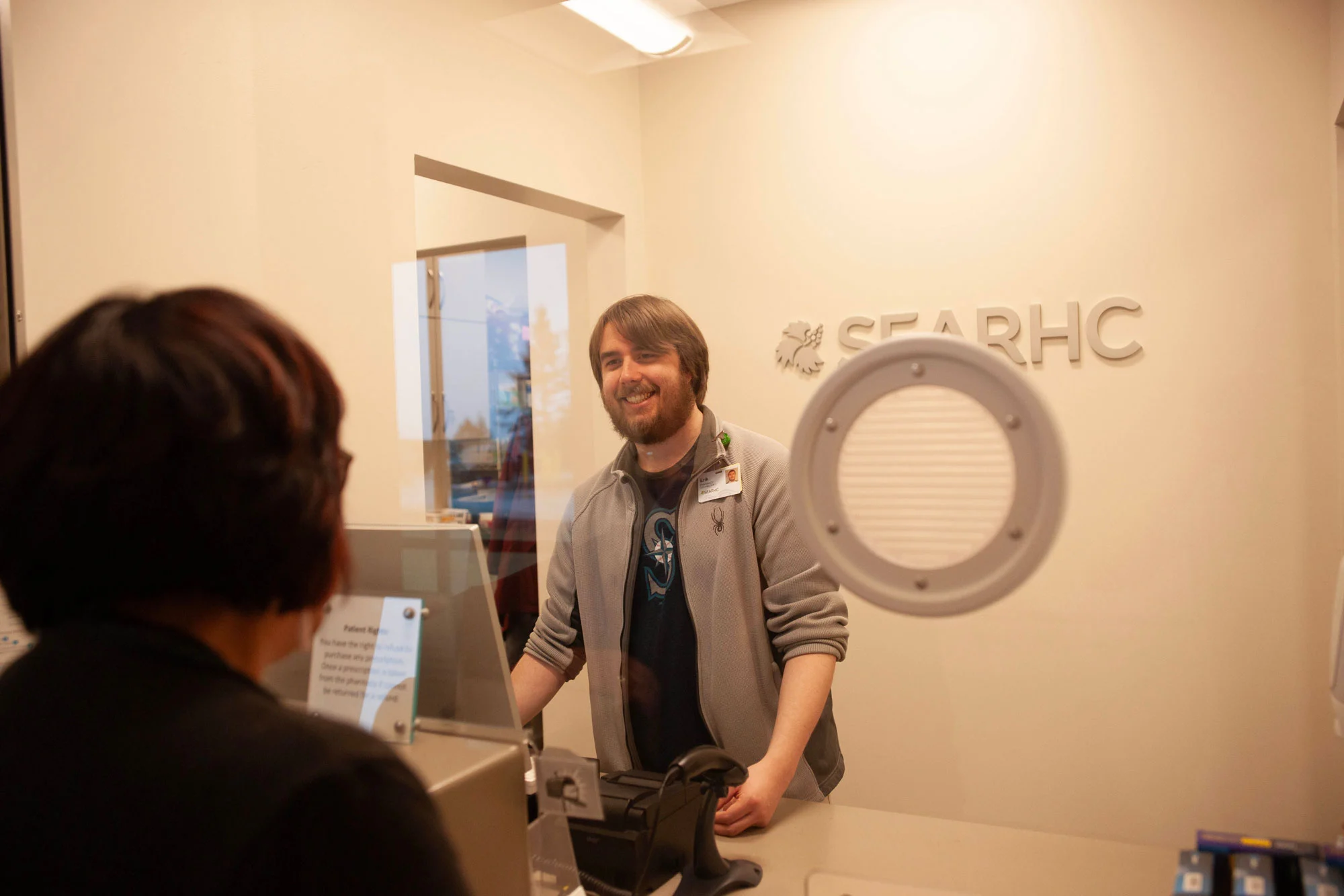 A smiling man with a beard behind a glass counter, facing the back of a person's head. A 'SEARHC' logo is on the wall behind him.