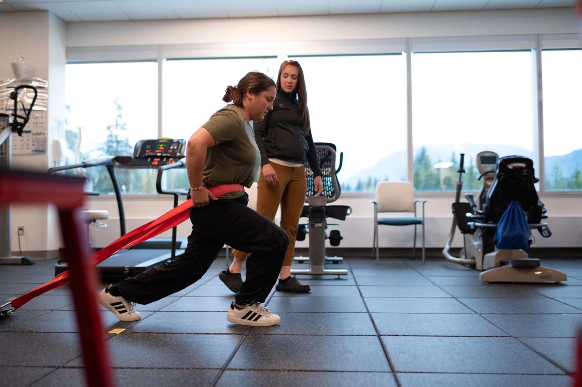 A woman performs a lunge with a red resistance band while a trainer observes in a bright gym with large windows.