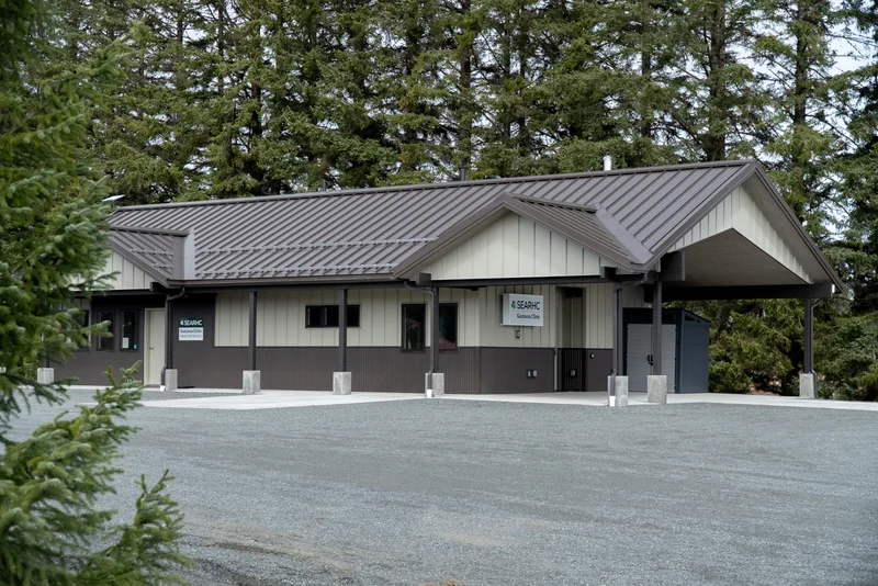 Exterior view of the SEARHC Gustavus Clinic, a modern building with a brown metal roof, light beige and dark brown siding, and a gravel parking area surrounded by evergreen trees.