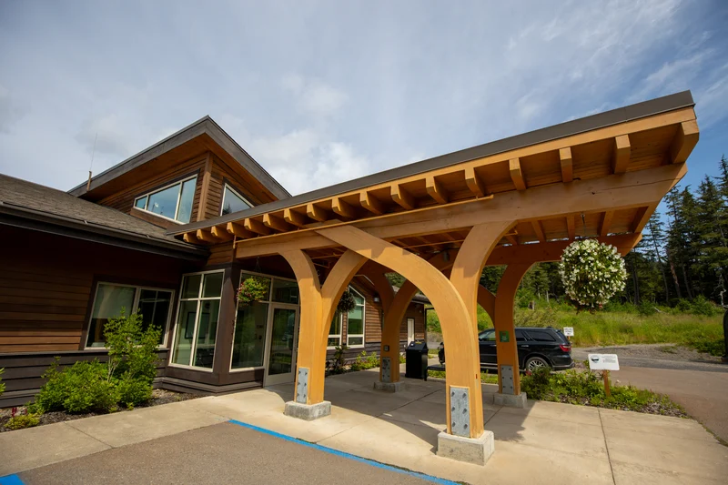 Modern building entrance with a large wooden canopy, hanging white flower baskets, and a concrete path. Green trees and a blue sky are in the background.