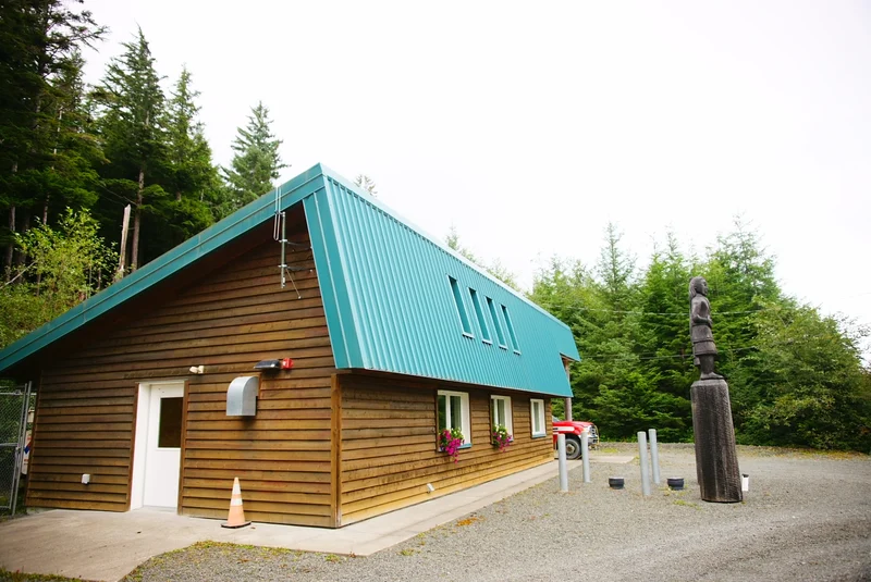 A wooden building with a teal metal roof and flower boxes, next to a tall wooden statue, surrounded by a gravel lot and dense forest.