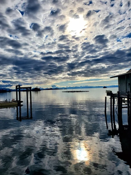 A calm body of water reflecting a cloudy sky with sunlight breaking through. Wooden structures are on the left and right, with distant land visible.