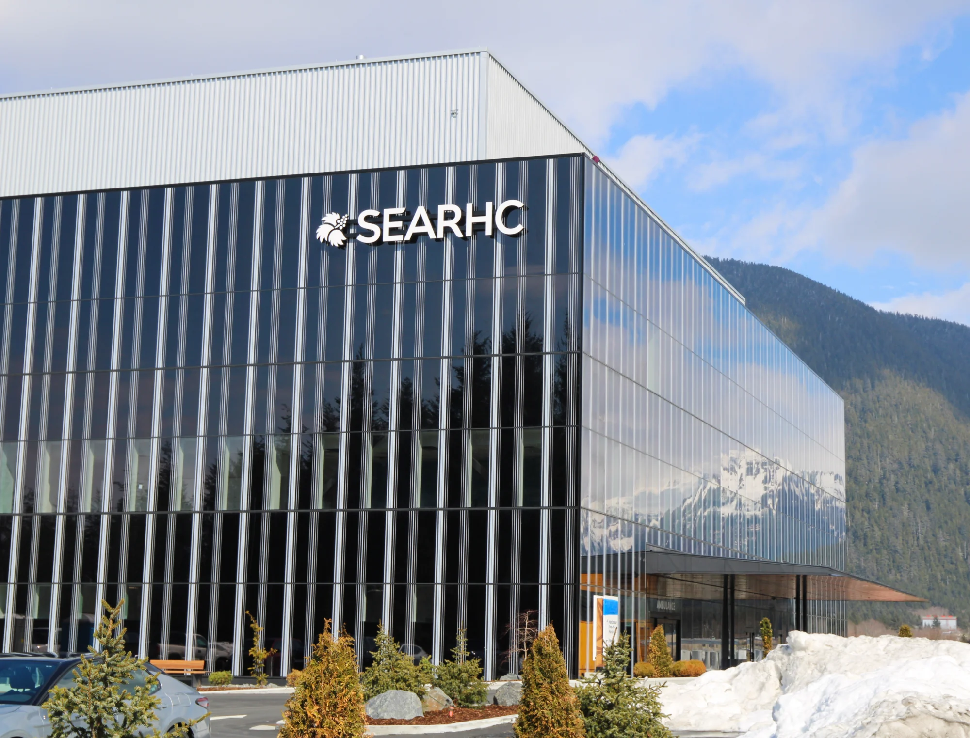 Exterior view of the modern SEARHC building with a glass facade reflecting mountains, under a blue sky, with trees and snow in the foreground.