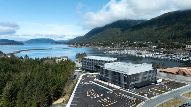 Aerial view of two modern buildings with a large parking lot, situated between a forest and a bay with a marina and forested mountains in the background.