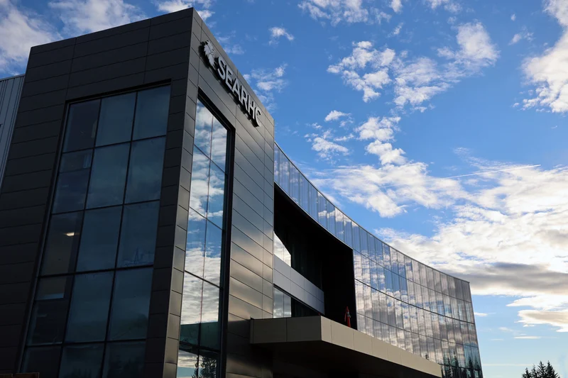 A modern building with a dark gray facade and extensive reflective glass windows. The SEARHC logo is visible on the left section. The glass reflects a blue sky with white clouds.