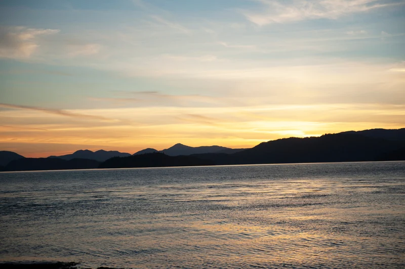 A colorful sunset or sunrise sky with orange and yellow hues above dark mountains and calm, rippling water.