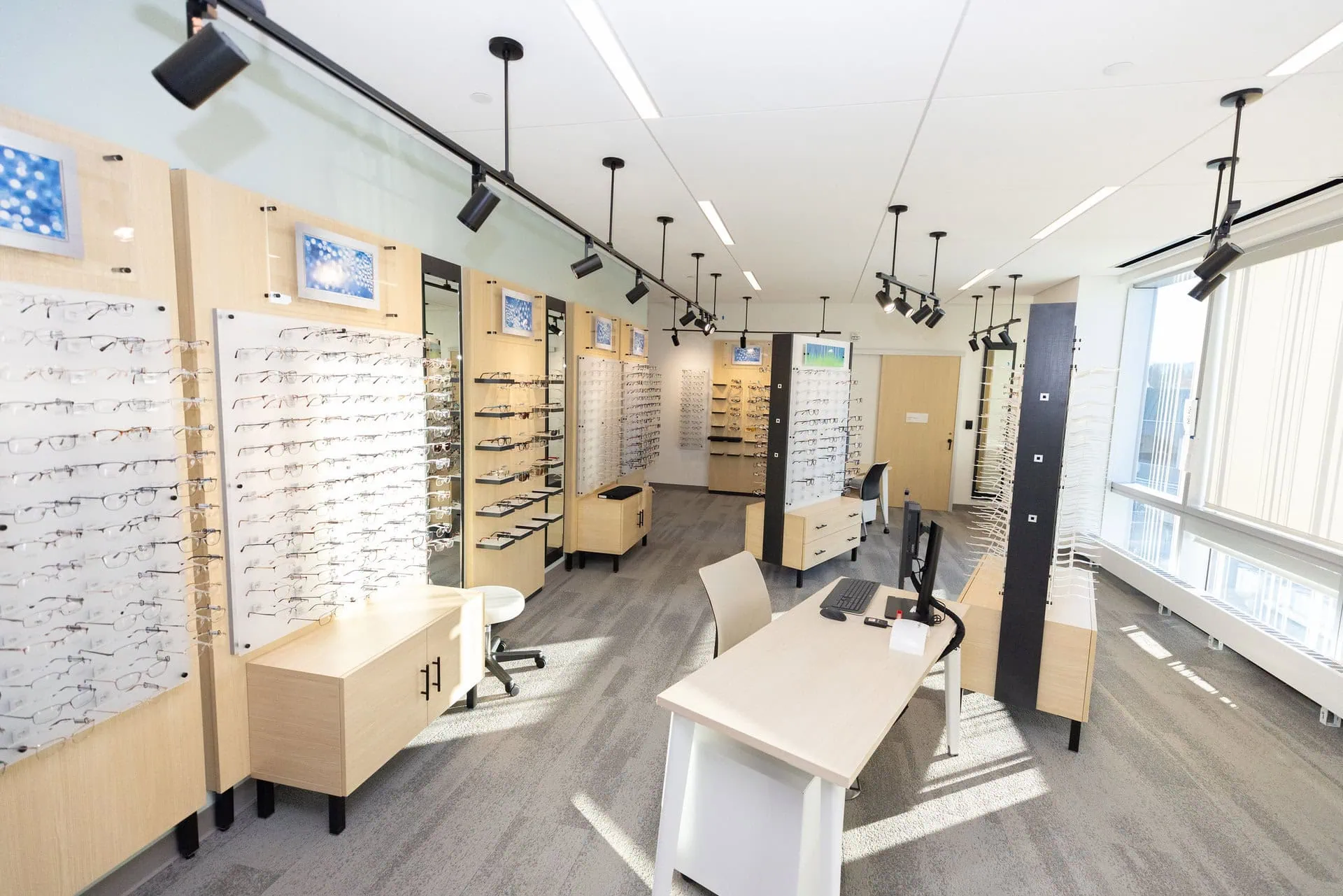 Interior view of a modern optical store with numerous eyeglasses displayed on wall panels and shelves, a central desk with a computer, and bright lighting.