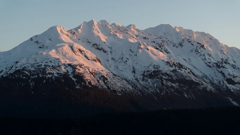 Snow-capped mountains at sunrise or sunset, with golden light on the peaks and dark trees below.