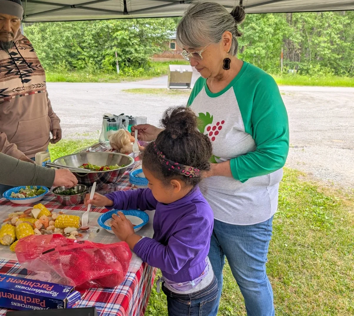 A young child and an adult woman serve themselves food from a table under a tent, with a man partially visible on the left. The table is covered with a plaid cloth and holds salad, corn, shrimp, and other items.