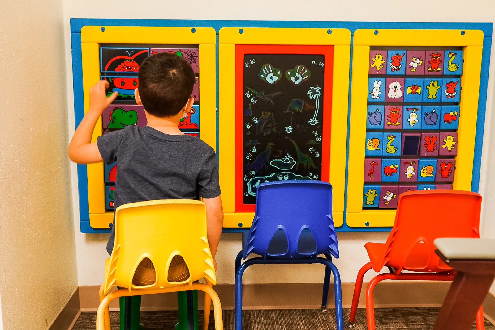 A young boy from behind, sitting on a yellow chair and playing with a colorful, wall-mounted activity board that has puzzles, a drawing surface, and animal matching squares.