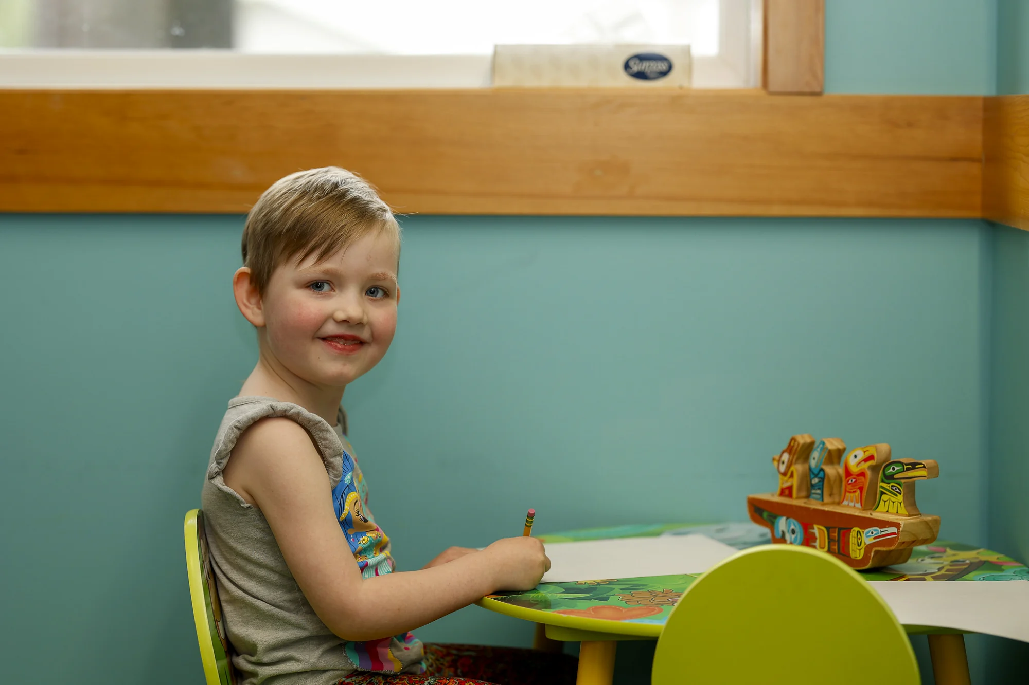 A smiling child with light hair sits at a colorful table, holding a pencil, with a wooden toy boat nearby.
