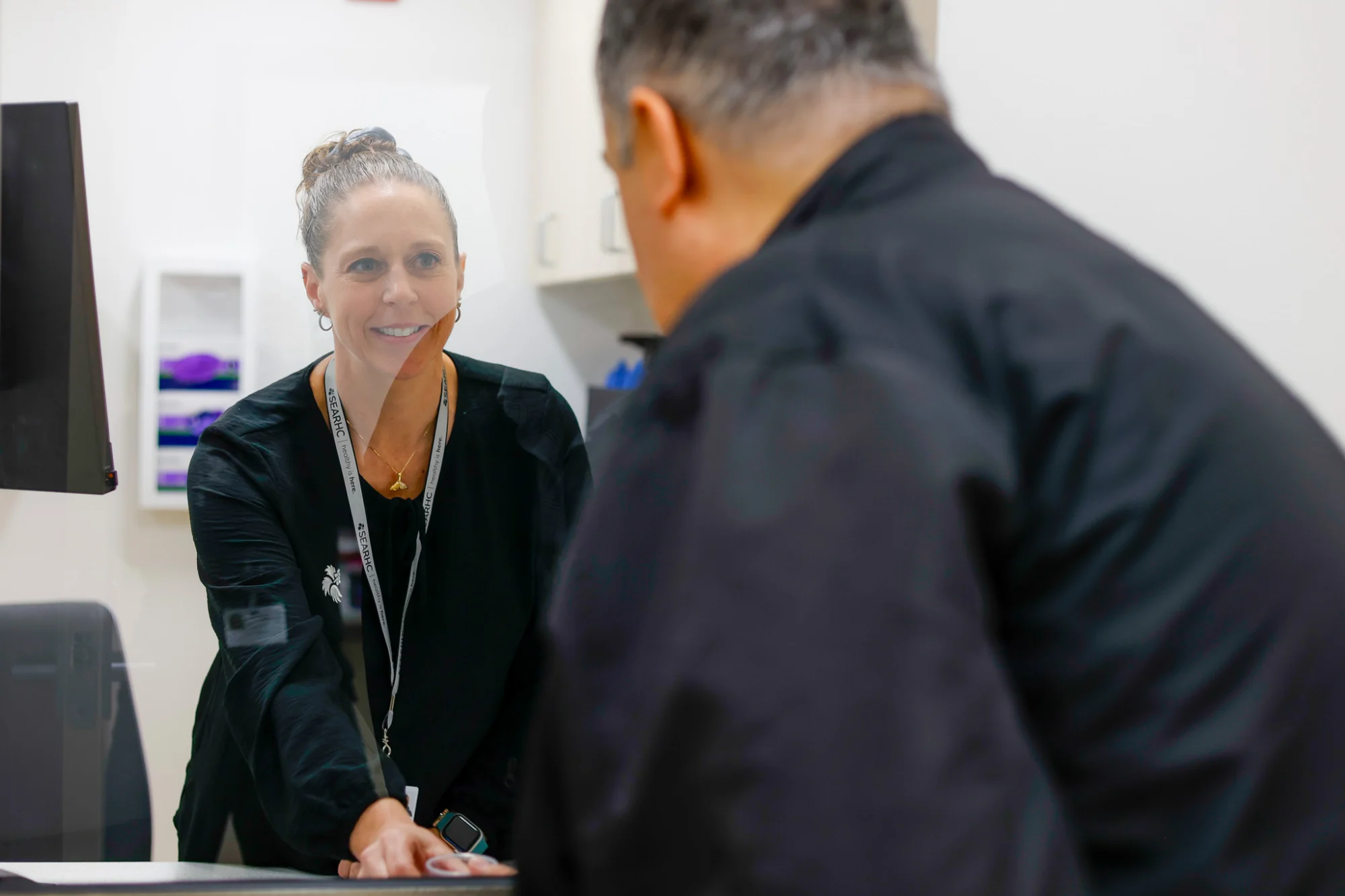 A smiling woman behind a clear barrier interacts with a man seen from behind at a reception desk. She wears a 'SEARHC' lanyard.
