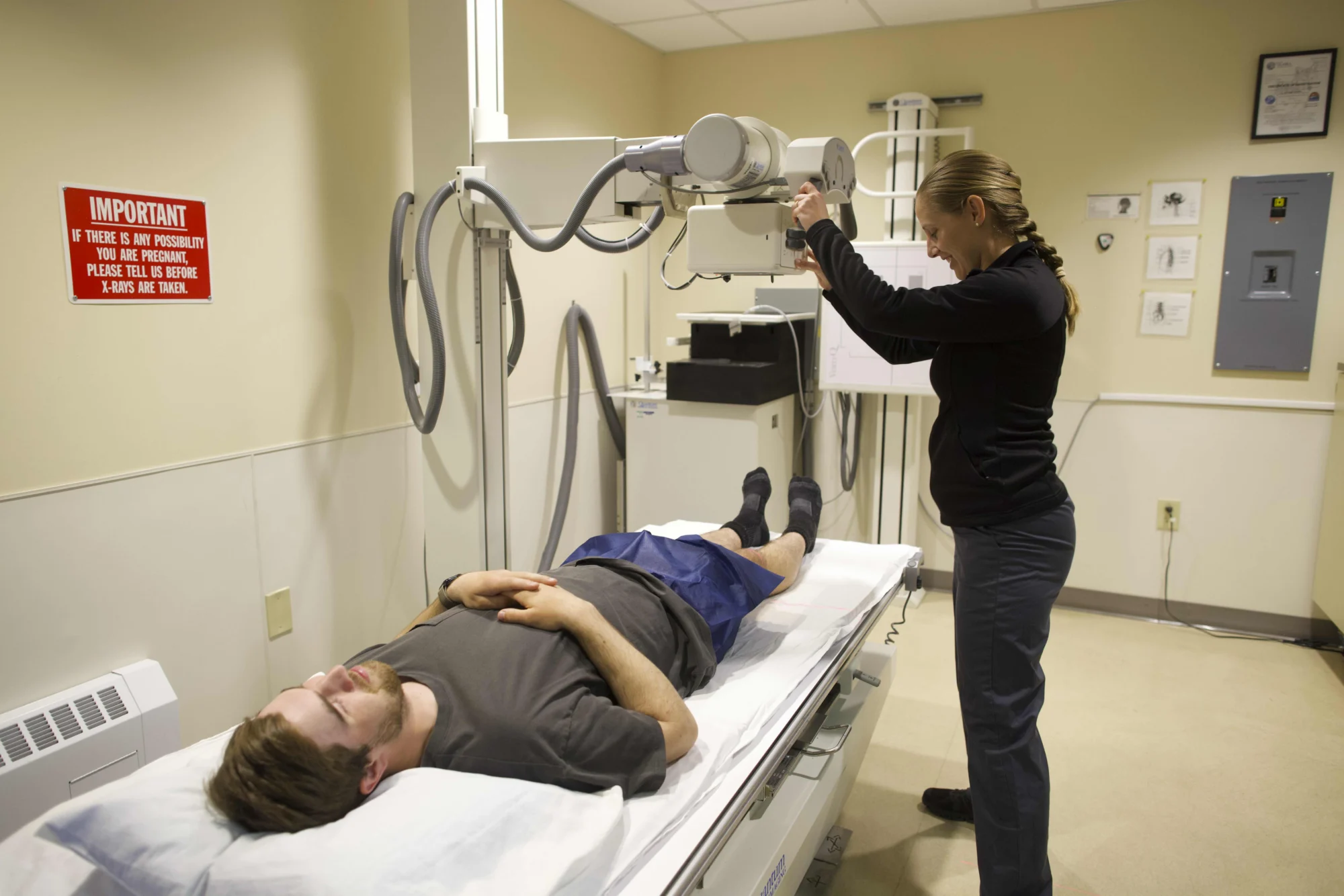 A female technologist adjusts an X-ray machine over a male patient lying on a table in a medical room.