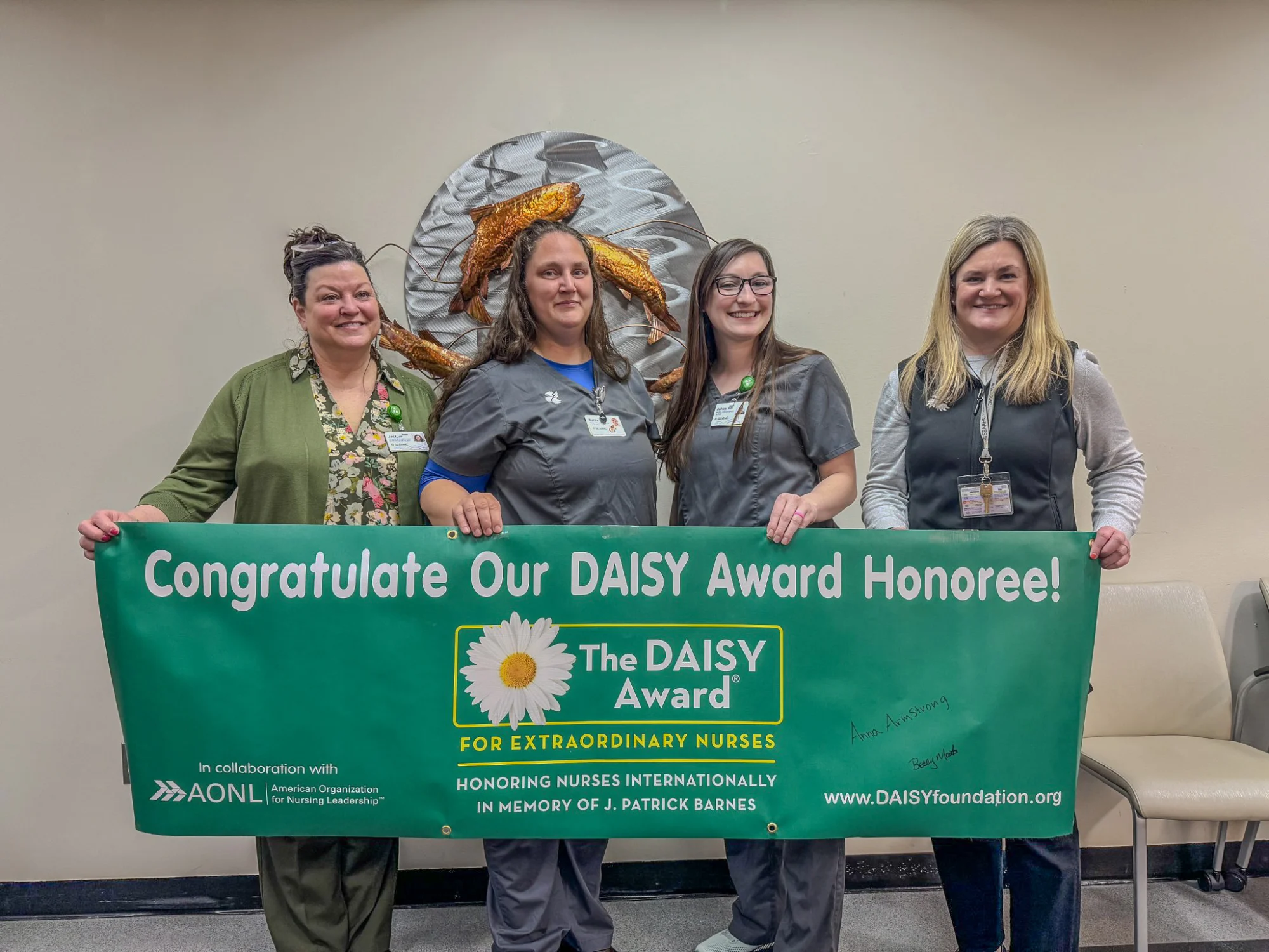 Four smiling women, likely nurses, hold a green banner celebrating a DAISY Award Honoree for extraordinary nursing.