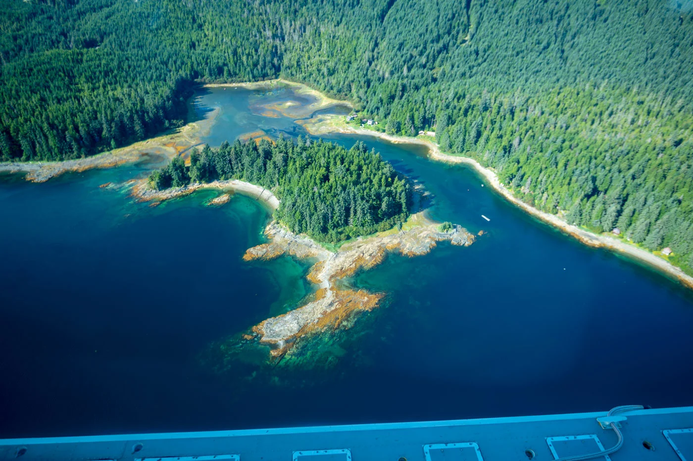 Aerial view of a dense green forest meeting a rugged coastline with deep blue and turquoise water, rocky outcrops, and a small tidal flat. A few small buildings are visible along the shore.