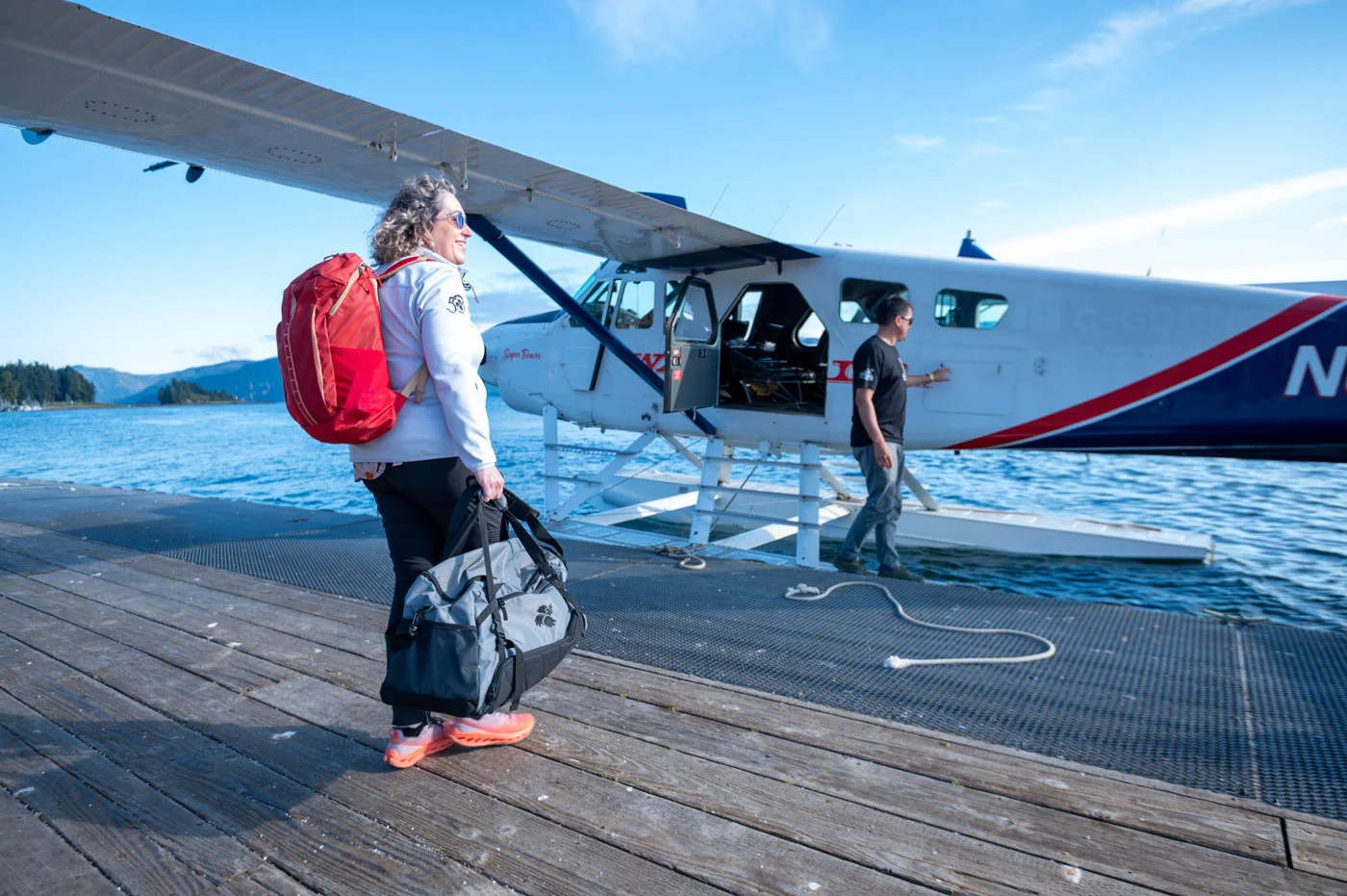 A woman with a red backpack and gray duffel bag stands on a wooden dock next to a white seaplane on blue water. A man is near the plane's open door.