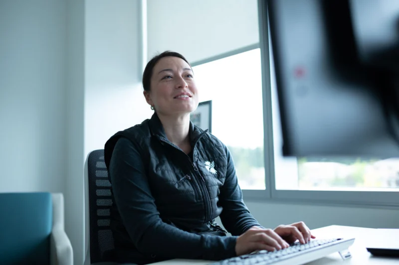 A woman in a black vest smiles while typing on a keyboard at a desk in a bright office.