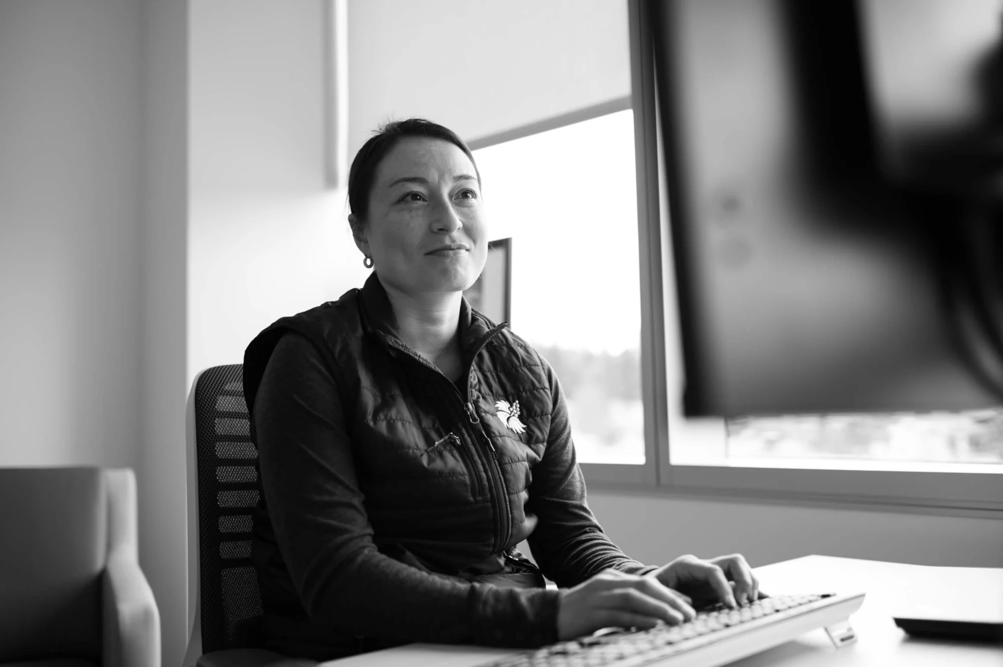 Black and white image of a woman with dark hair and freckles, wearing a dark vest, sitting at a desk with her hands on a keyboard, looking up and to the right with a slight smile. A window is behind her.