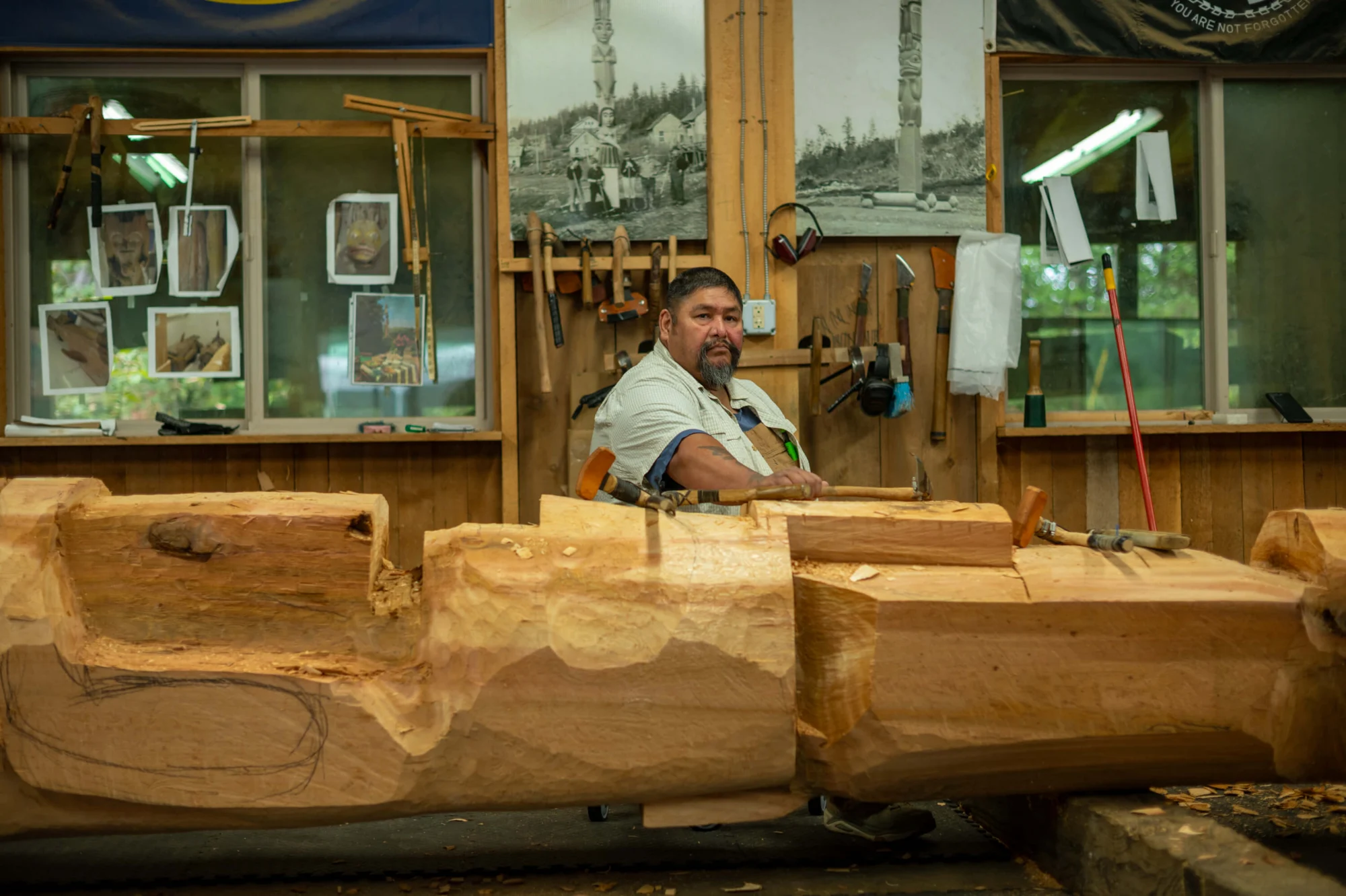 A man with a beard carves a large wooden log in a workshop, holding an adze. Tools and reference photos hang on the wooden walls behind him.