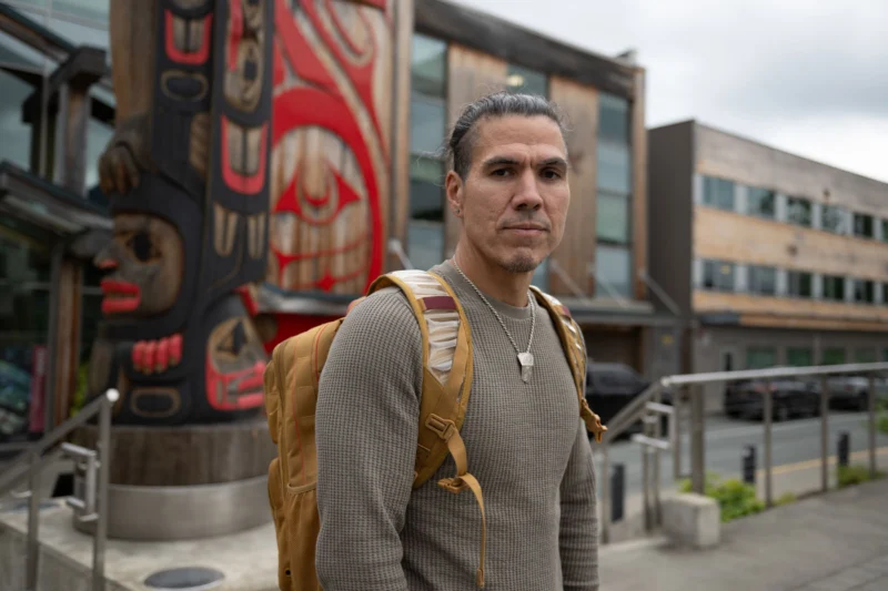 A man with a backpack stands in front of a carved wooden totem pole and modern buildings.