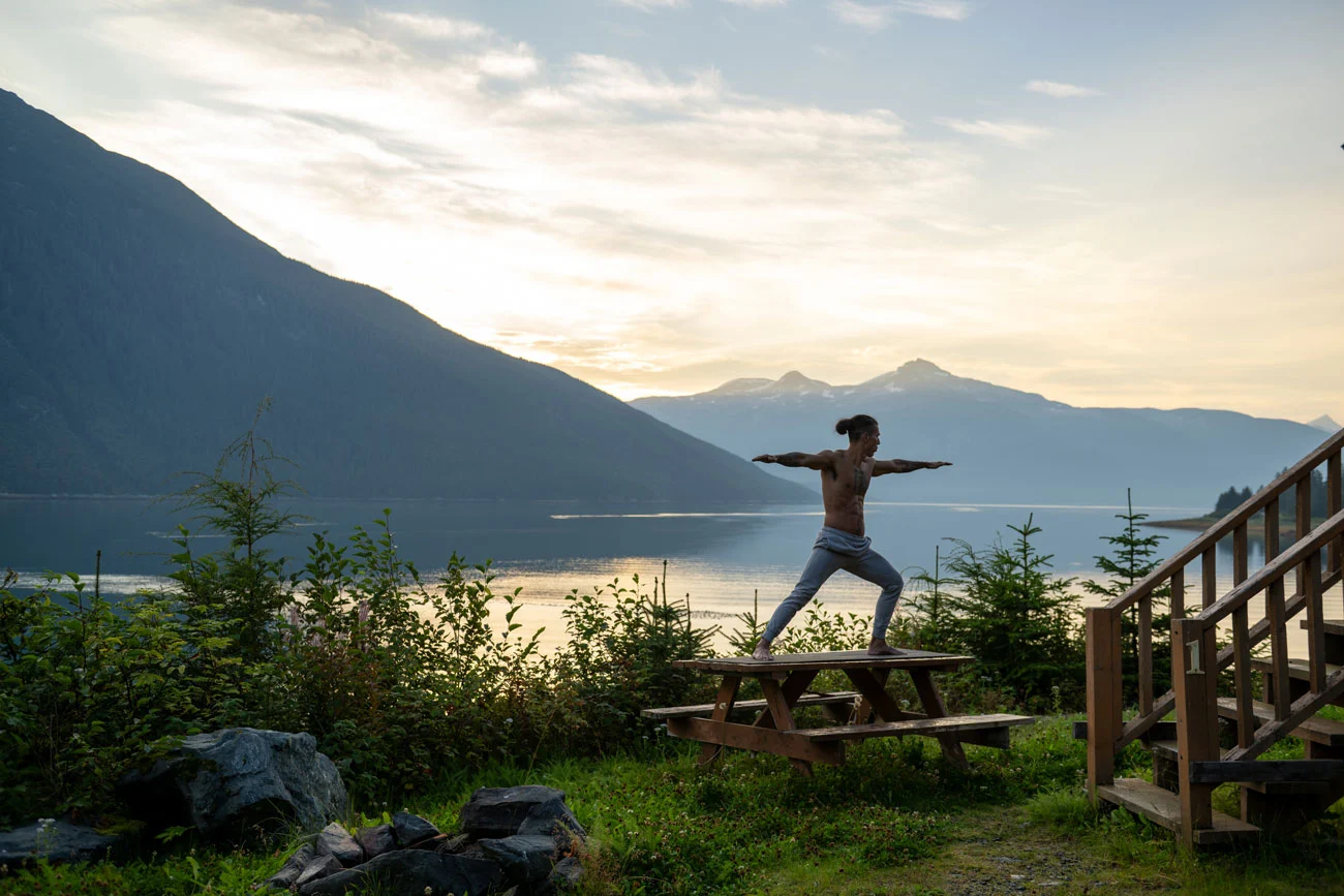 A man in Warrior II yoga pose stands on a picnic table by a lake surrounded by mountains under a hazy sky.